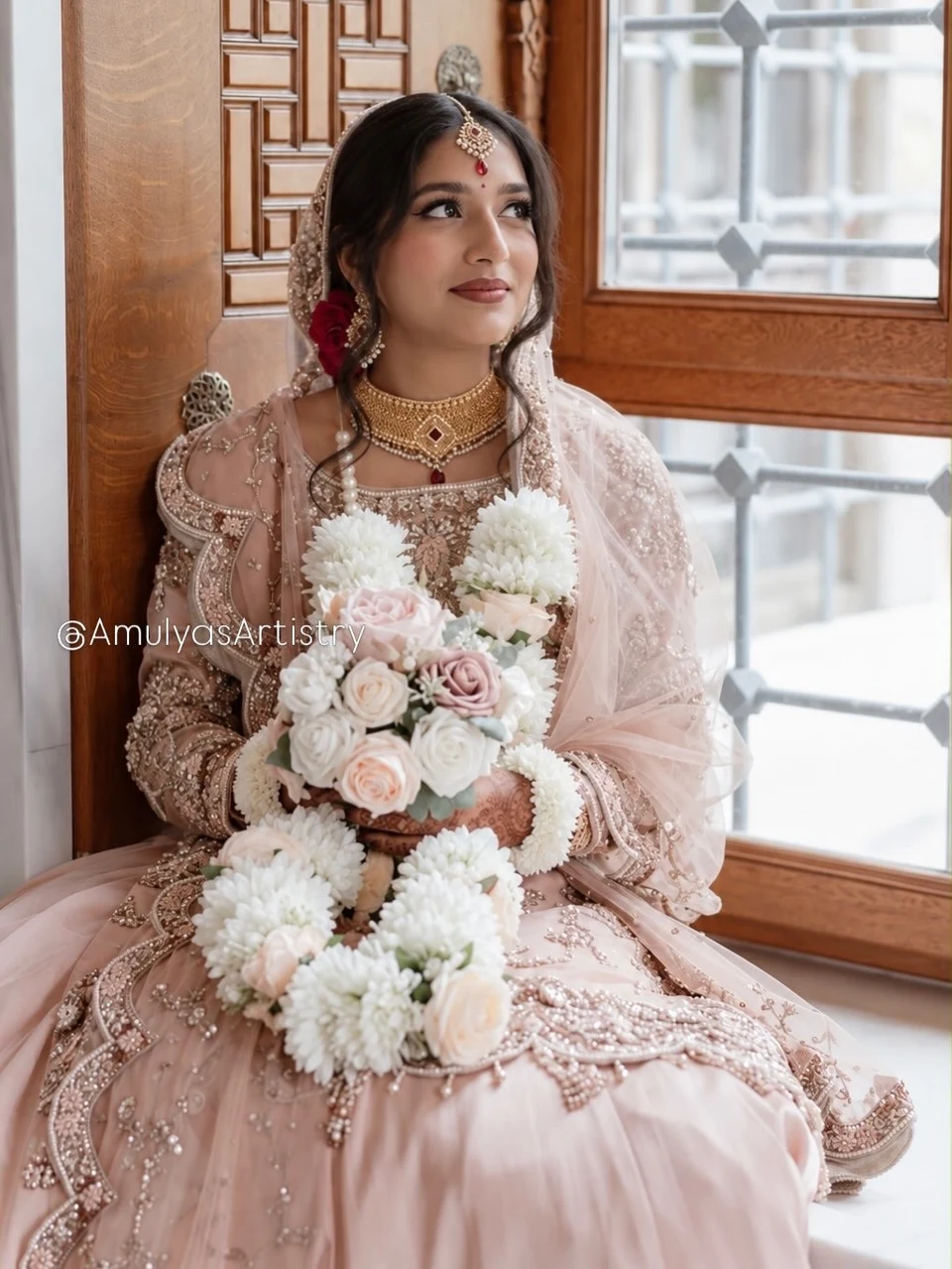 Bride in a blush outfit holding a bouquet of pale roses by a window