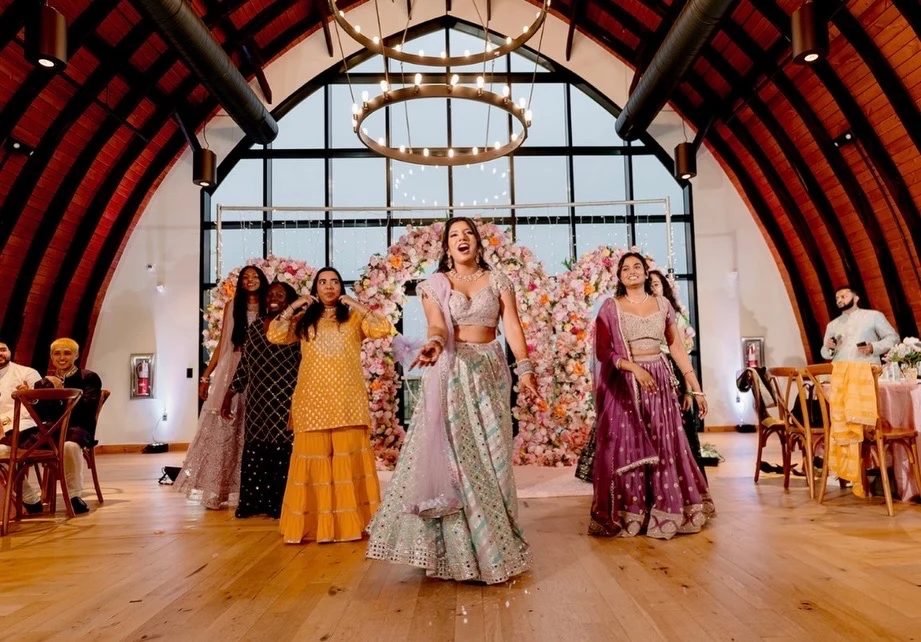 Group dancing in colorful occasion outfits beneath a timber ceiling and floral decorations