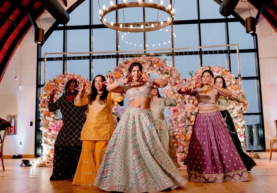 Group in colorful embroidered occasion outfits dancing in front of pink floral decorations