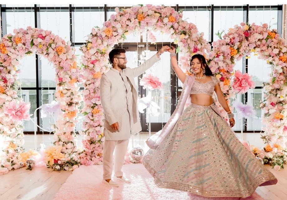 Couple twirling beside pink floral decor, with a flowing pastel embroidered lehenga