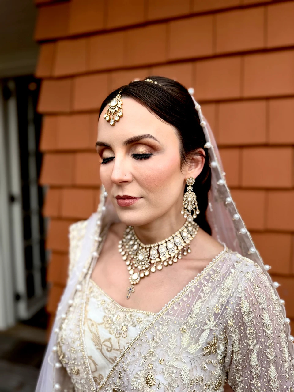 Close bridal portrait showing shimmering eye makeup, softly defined lips and pearl jewelry beneath a pale veil